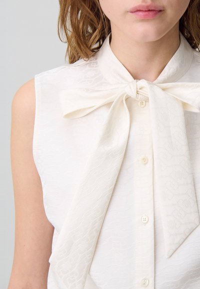 Blusa de tirantes blanca con cuello de lazo, que presenta un patrón texturizado, cierre de botones y una tela suave.