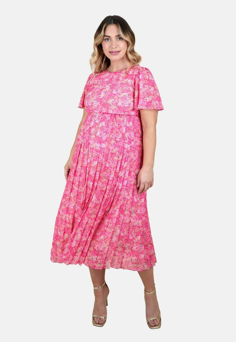 Nő áll, rózsaszín virágmintás midi ruhában, rövid ujjúval és arany bokapántos magassarkú cipőben, egyszerű világos háttér előtt.