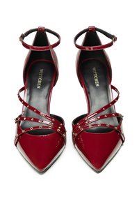 Scarpe col tacco alto in vernice rossa con punte affusolate, cinturini alla caviglia e fasce decorative borchiate sul davanti. Logo del produttore visibile all'interno.