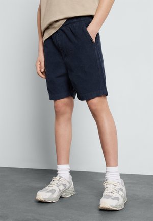 Barn iført marineblå kordbukser, beige t-shirt, hvide crew-sokker og hvide sportssko står på gråt gulv.