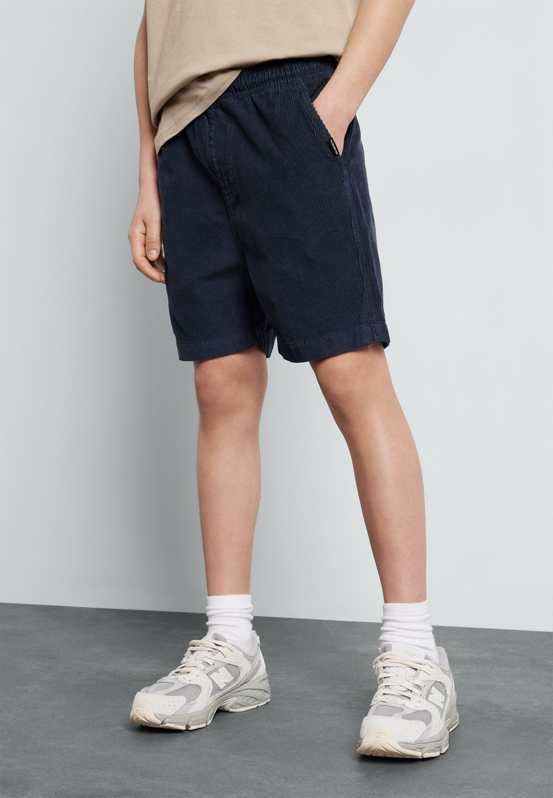 Enfant portant un short en velours côtelé bleu marine, un t-shirt beige, des chaussettes blanches et des baskets blanches, debout sur un sol gris.