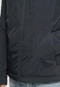 Dunkle, leichte Jacke mit glatter, glänzender Textur; ausgestattet mit einem vollständigen Frontreißverschluss und Seitentaschen; im minimalistischen Stil gestaltet.