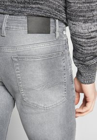 Sivé denímové rifle s textúrovaným povrchom a čiernym koženým štítkom na páse. Obsahujú zadné vrecká s decentnými detailmi prešitia.