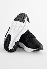 Czarno-białe sportowe sneakersy z teksturowanymi gumowymi podeszwami, jeden but oparty na drugim, prezentowane na białym tle.