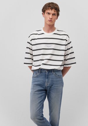 Gestreepte T-shirt met korte mouwen in wit en zwart, met de tekst "DE GEEST VAN DE STAD" op de borst, gecombineerd met lichtblauwe jeans.