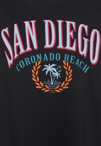Svart t-shirt med "SAN DIEGO" i stora vita och rosa bokstäver, "CORONADO BEACH" i turkost, och en grafik av palmer och vågor i orange.