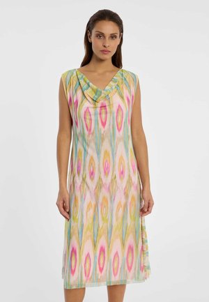 Frau trägt ärmelloses, knielanges Kleid mit mehrfarbigen abstrakten vertikalen Mustern in Pink, Gelb, Blau und Grün auf hellem Hintergrund.