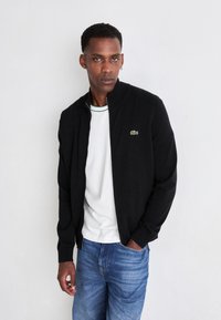 Man wearing black zip-up jacket, white t-shirt, and blue jeans, standing against plain white background with one hand in pocket.