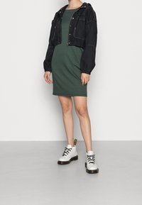 Lyhyt musta takki, jossa edessä vetoketju ja kaksi taskua, yhdistetty vihreään polvimittaiseen mekkoon ja valkoisiin nauhattomiin nilkkasaappaisiin.