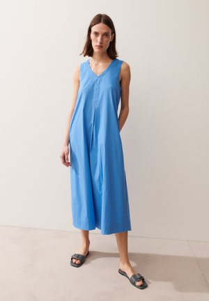 Vrouw staat tegen een witte muur, draagt een mouwloze blauwe midi-jurk en zwarte instapsandalen.