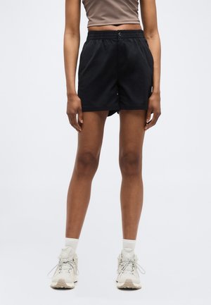 Persoană purtând pantaloni scurți negri până la mijlocul coapselor, top scurt bej, șosete albe și pantofi sport albi, stând pe un fundal simplu și deschis la culoare.