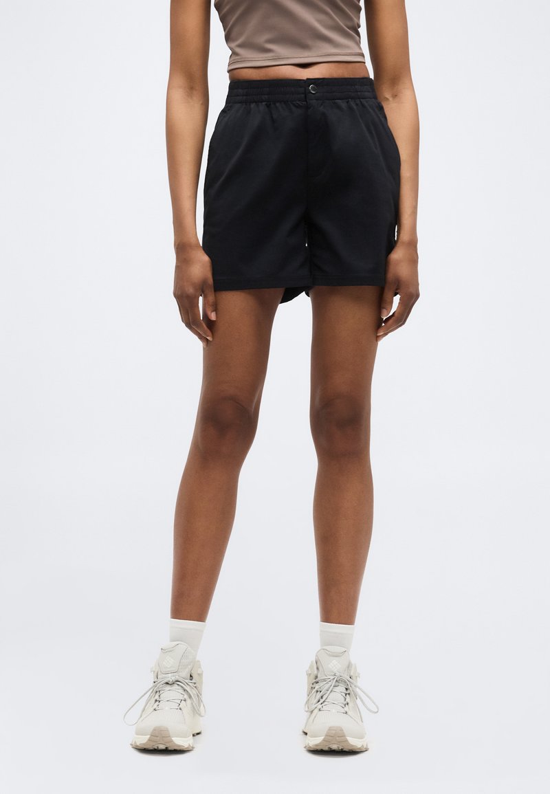 Άτομο που φοράει μαύρα σορτς μέχρι το μέσο του μηρού, μπεζ κοντό τοπ, λευκές κάλτσες και λευκά αθλητικά παπούτσια, στέκεται μπροστά σε απλό φωτεινό φόντο.