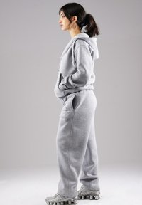 Young woman with dark ponytail wearing large hoop earrings, gray hoodie, matching sweatpants, and silver sneakers, standing in profile against gray background.
