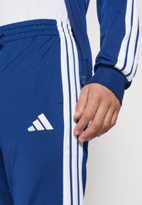 Modre atletske trenerke z belimi treh črtastimi detaile in vezenim logotipom. Tkanina je gladka z blagim sijajem.