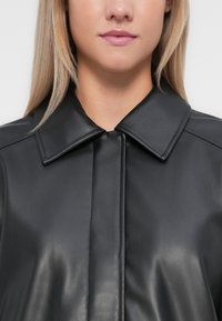 Veste en cuir noir avec une texture lisse, grand col et fermeture à boutons. Présente des coutures discrètes le long des coutures.