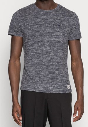 Homme portant un t-shirt gris chiné à manches courtes et un pantalon noir, debout les bras détendus le long du corps sur un fond uni.