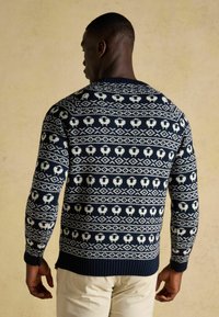 Blauer Strickpullover mit einem weißen Muster aus Herzen und geometrischen Formen, gerippten Bündchen und Saum sowie einem runden Kragen.
