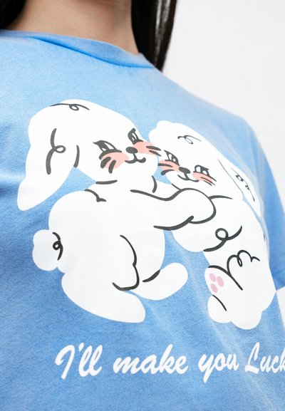 Μπλε βαμβακερό t-shirt με τρία καρτούν λαγουδάκια σε λευκό χρώμα με ροζ λεπτομέρειες, παιχνιδιάρικες εκφράσεις και το κείμενο «Θα σε κάνω τυχερό» κάτω από αυτά.