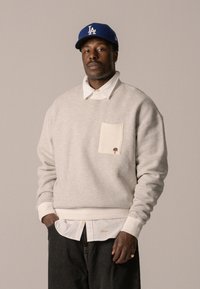 Grauer Kapuzenpullover mit cremefarbener Tasche, gesticktem Baumdetail. Über einem weißen Hemd mit Kragen getragen. Dunkle Jeans, cremefarbene gerippte Bündchen an den Handgelenken. Blauer Baseballcap.