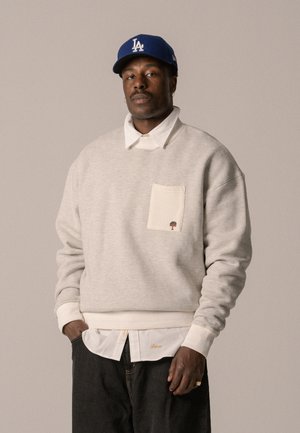 Grijze sweatshirt met een crèmekleurige zak en geborduurd boomdetail. Gelaagd over een witte blouse. Donkere spijkerbroek, crèmekleurige geribde manchetten bij de polsen. Blauwe pet.