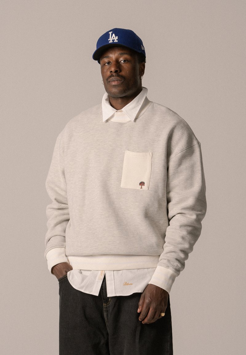 Grauer Kapuzenpullover mit cremefarbener Tasche, gesticktem Baumdetail. Über einem weißen Hemd mit Kragen getragen. Dunkle Jeans, cremefarbene gerippte Bündchen an den Handgelenken. Blauer Baseballcap.