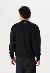 Sudadera negra con cuello redondo, puños y dobladillo acanalados, con un pequeño logo verde en la parte superior de la espalda. Textura suave, corte relajado.