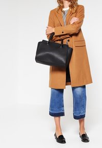 Sort læderhåndtaske med struktureret form, dobbelte toppehåndtag og glat tekstur, parret med en tan-frakke og cropped blå jeans.