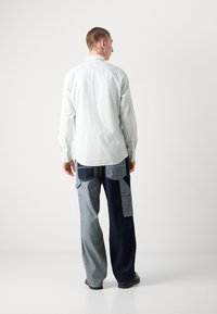 Camisa de manga larga verde claro con puños abotonados, combinada con pantalones de mezclilla de patchwork en diferentes tonos de azul, que cuentan con grandes bolsillos.