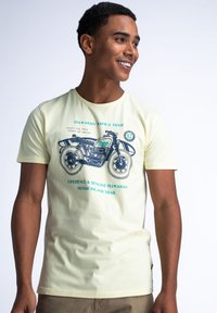 Světle žluté tričko s krátkým rukávem s grafikou motocyklu a modrým textem. Měkký bavlněný materiál, kulatý výstřih, volný střih.