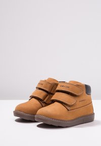 Braune wasserdichte High-Top-Schuhe mit doppelten Velcro-Riemen, glatter Wildleder-Oberfläche, Gummisohle und dezenten Details an den Seiten.