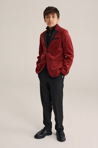 Blazer en velours rouge avec revers et poches noirs, associé à un pantalon noir et des chaussures noires. Texture lisse, coupe ajustée et aspect sur mesure.