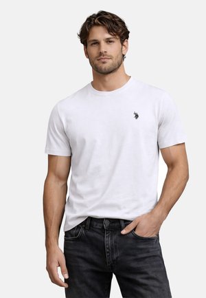 Man with light skin and brown hair wearing a white short-sleeve t-shirt with a small logo on the chest and black jeans, posing with hand in pocket.