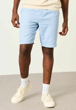 Mann trägt hellblaue Shorts, weiße Socken und cremefarbene Sneakers und steht auf einem grauen Boden vor einem hellen Hintergrund.
