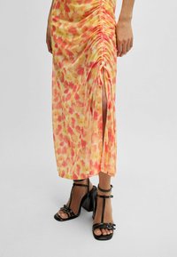 Yellow-orange midi dress with a floral pattern, ruched detailing, and side slit. Paired with black strappy high-heeled sandals.