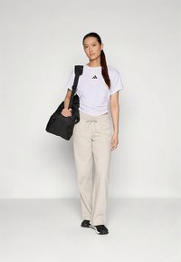 Ung kvinna i vit Adidas-t-shirt och ljusbeige byxor, bär en svart duffelväska över ena axeln, står mot en enfärgad bakgrund.