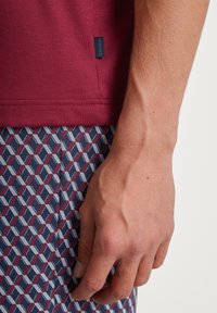 Bordeauxrotes Baumwollhemd mit einem seitlichen Etikett, kombiniert mit blau-rot gemusterten Shorts, die geometrische Formen zeigen. Nahaufnahme einer Hand, die auf den Shorts ruht.