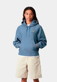 Sudadera azul claro con un bolsillo canguro frontal, cordones ajustables y un pequeño logo. Combinada con pantalones cortos beiges, que cuentan con una tela texturizada.