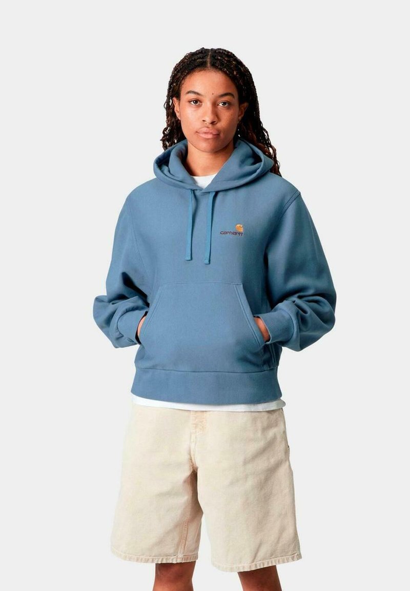 Sudadera azul claro con un bolsillo canguro frontal, cordones ajustables y un pequeño logo. Combinada con pantalones cortos beiges, que cuentan con una tela texturizada.