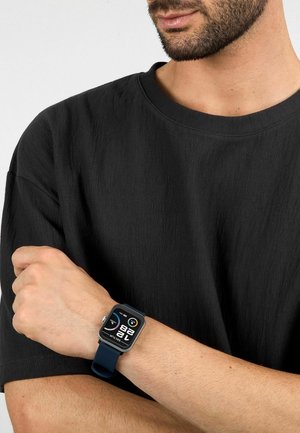 Smartwatch med rektangulært urværk, sort display og digital aflæsning. Har et navyblåt silikonebånd, båret på et håndled med lys hudtone.
