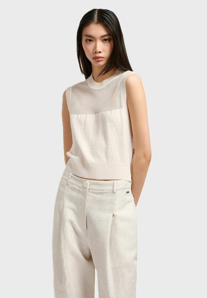 Top senza maniche in maglia beige chiaro con un design texturizzato e un pannello superiore in rete trasparente, abbinato a pantaloni a sigaretta crema con un motivo geometrico.