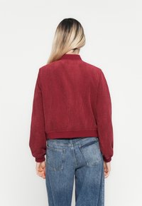 Person with long blonde hair wearing a red ribbed jacket and blue jeans, standing facing a plain white background.
