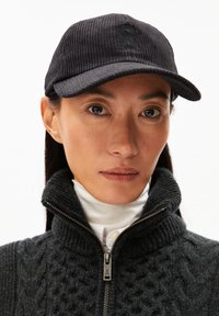 Zwarte corduroy pet met een gebogen rand en subtiel logo. Model draagt een donkere trui met hoge kraag en ritsdetail. Gladde huidtextuur zichtbaar.