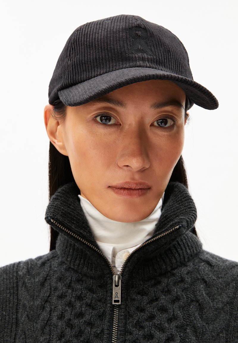 Zwarte corduroy pet met een gebogen rand en subtiel logo. Model draagt een donkere trui met hoge kraag en ritsdetail. Gladde huidtextuur zichtbaar.