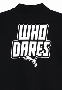 Polo shirt nera con testo bianco audace "WHO DARES" e logo grafico Puma. Tessuto liscio con un design del colletto classico.