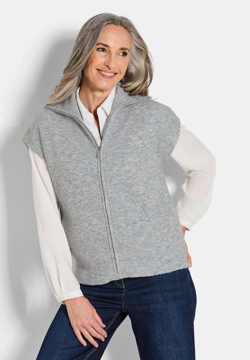Femme aux cheveux gris portant un gilet en tricot zippé gris clair sur une blouse blanche et un jean bleu foncé, souriante et posant avec la main sur la hanche.