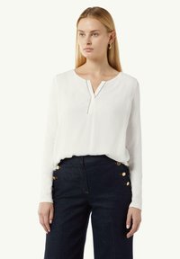 Weißes langärmliges Oberteil mit V-Ausschnitt und Knopfdetail, kombiniert mit dunkelblauen Jeans, die an den Seiten goldene Knopf-Details aufweisen.
