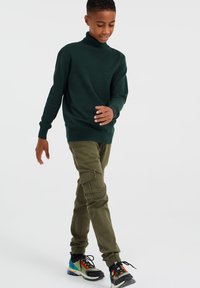 Zeleni pulover s turtleneck ovratnikom, olivno zeleni cargo hlače z stranskimi žepi in barvni superge z različnimi vzorci hodijo po enotnem ozadju.