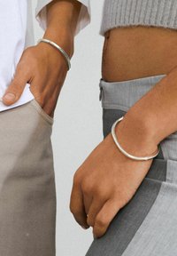 Two individuals wearing silver metal bangles on their wrists. One bangle is curved, while the other is round and smooth.