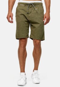 Shorts verts olive en tissu texturé avec une taille à cordon de serrage réglable, dotés d'une poche zippée et d'un ourlet roulé. Portés avec des baskets blanches.