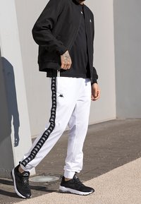 Giacca bomber nera, t-shirt nera, pantaloni jogger bianchi con strisce nere ai lati e scarpe da ginnastica nere. Texture lisce e colori a contrasto.